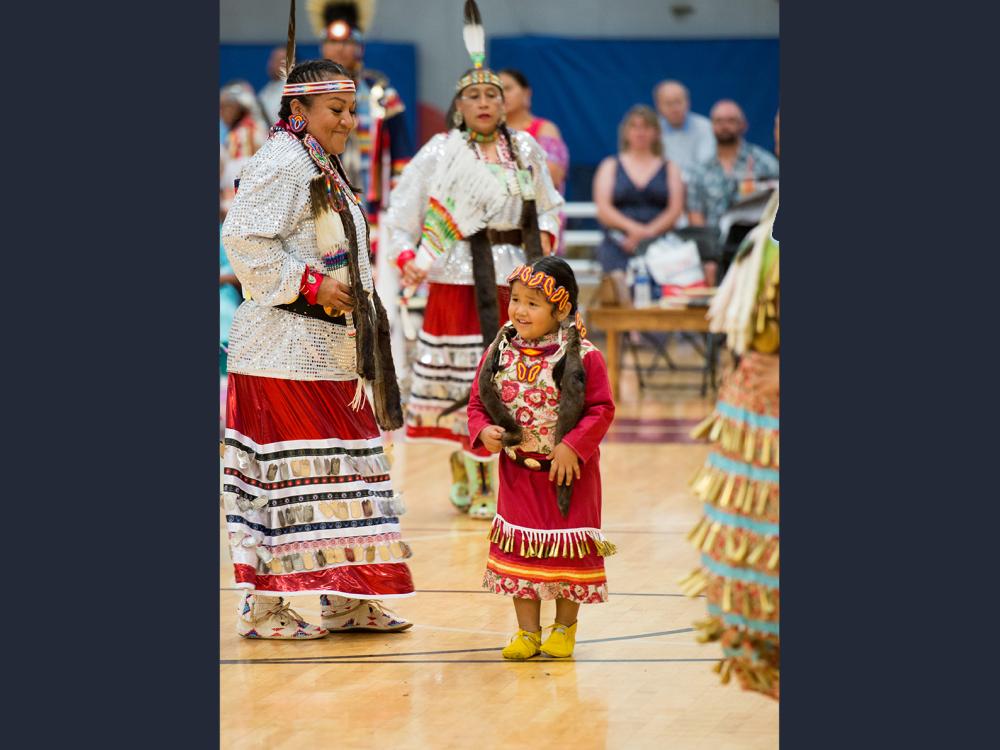 A child in traditional powwow clothing dances with an adult in a group