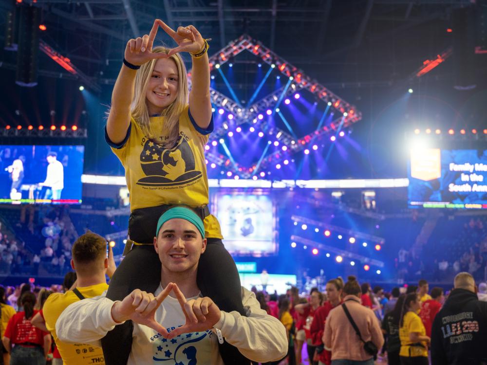 A female student sits on a male student's shoulders while both making diamond shapes with their fingers