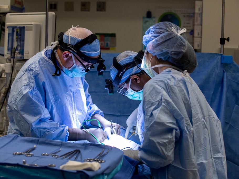 Three people dressed in full scrubs, caps and gloves work on a patient in an operating room. Several surgical tools are laying on a tray in the foreground.