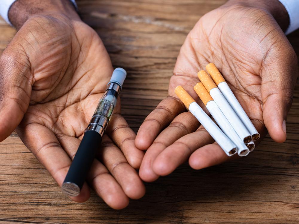 Two hands with the palms facing up hold items. The right hand contains an electronic cigarette. The left holds five cigarettes.