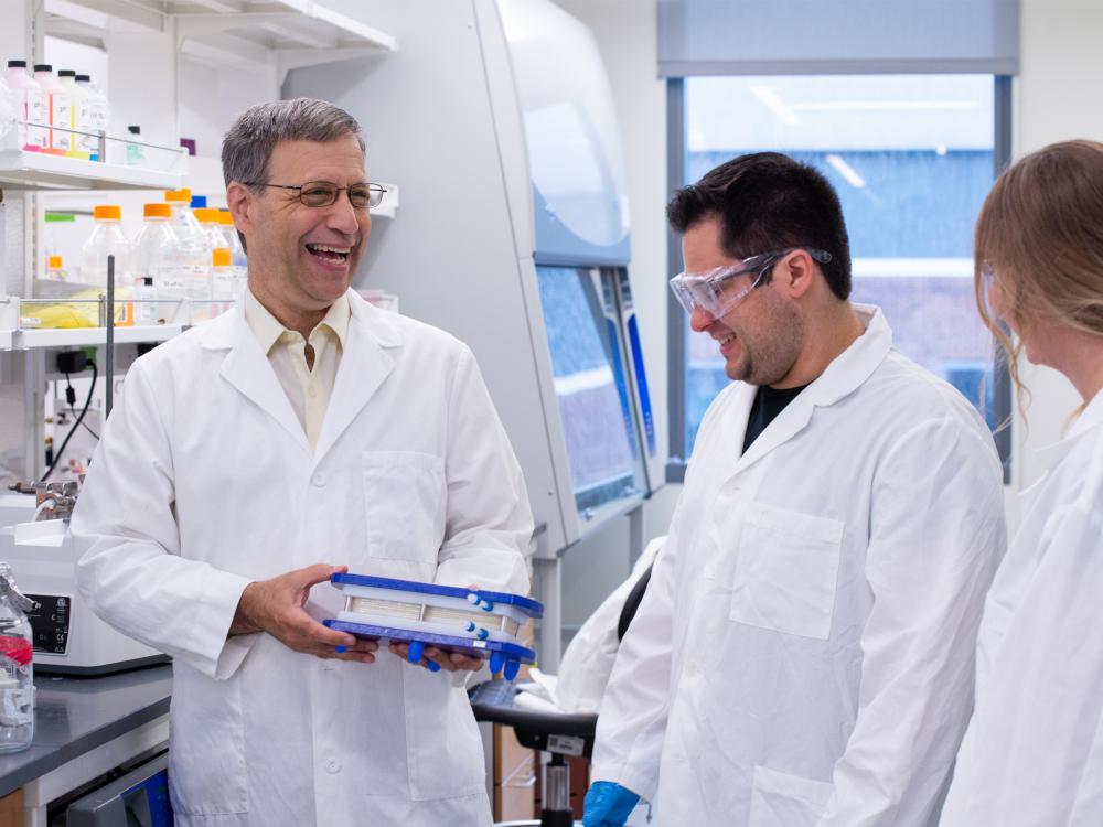 Three individuals in lab coats and protective glasses converse and smile in a lab setting.