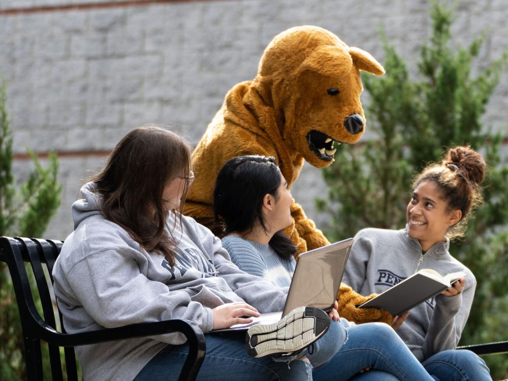 Three female students on a bench outdoors with Nittany Lion mascot leaning over them pointing to a book.