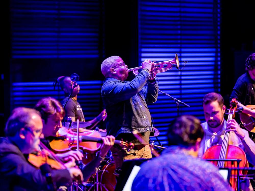 A Black man stands and plays a trumpet while a string quartet and jazz band perform nearby.