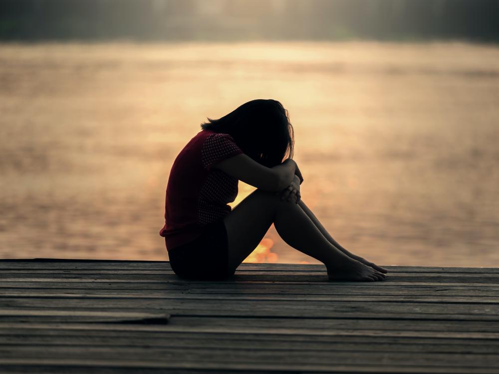 A young person with their head in their arms, silhouetted by a sunset over the ocean