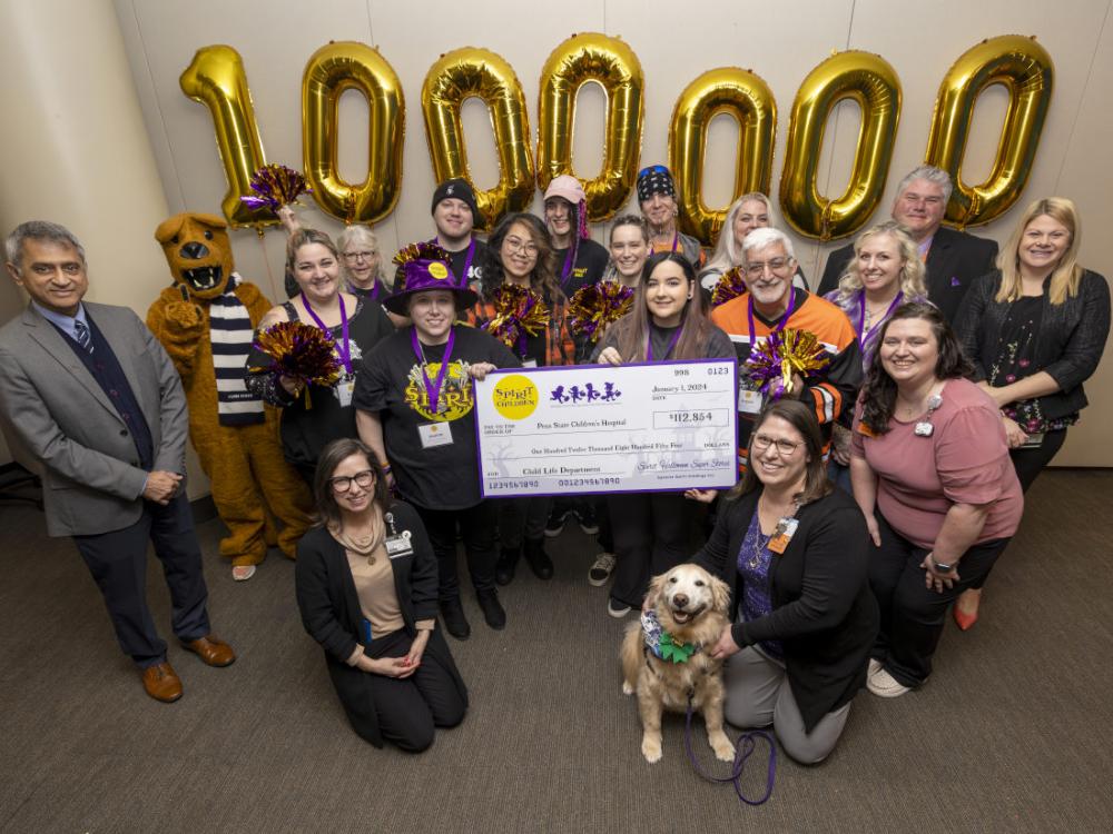 Eighteen people – along with a dog and a costumed Nittany Lion – pose with a check made out to Penn State Health Children’s Hospital in the amount of $112,854, from Spirit of Children. Large gold inflatable numbers spelling out “1,000,000” are on the wall directly behind them.