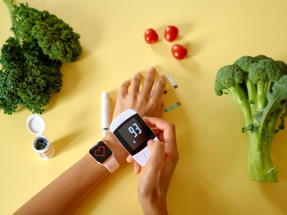 Close-up of an individual using a digital blood glucose monitor on themselves. Broccoli and tomatoes are on same table where the person is resting their arms.