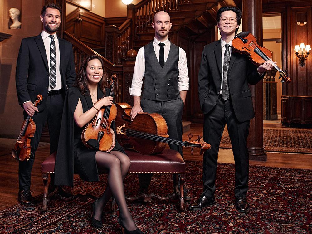 Three men stand with their string instruments while an Asian woman sits on a leather armchair cradling her instrument.