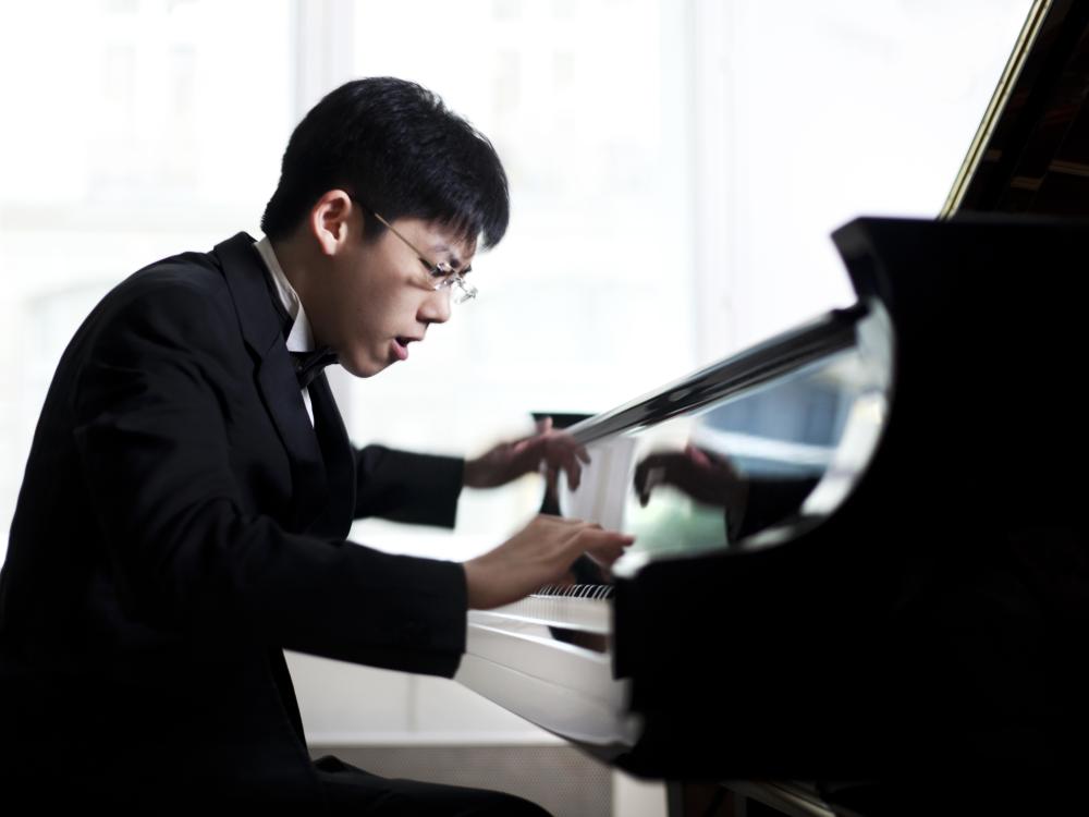 An Asian man sits at a piano and plays the keys with intensity.