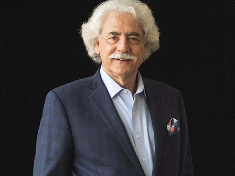 Older man with light grey hair wearing a navy blue suit with his hands in his pockets