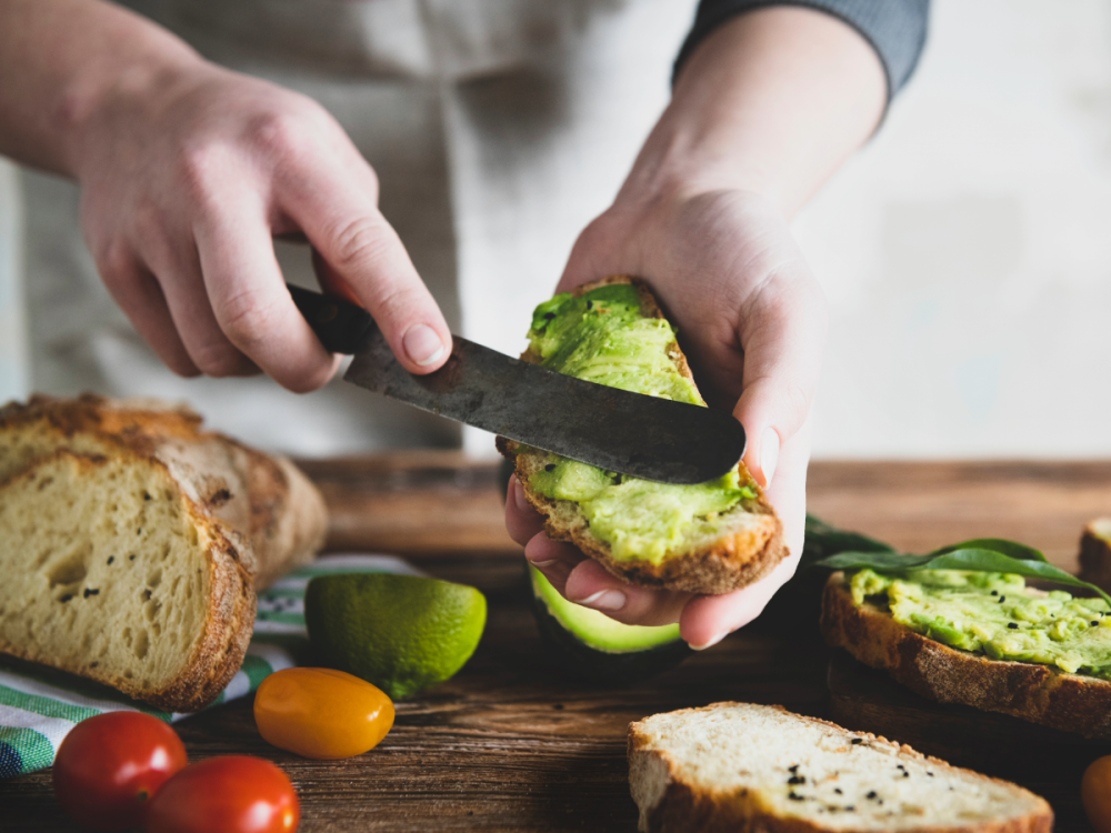 A man holds a piece of bread and uses a knife to spread pureed avocado on the bread. The table has a loaf of bread, tomatoes, and other slices of avocado and bread.