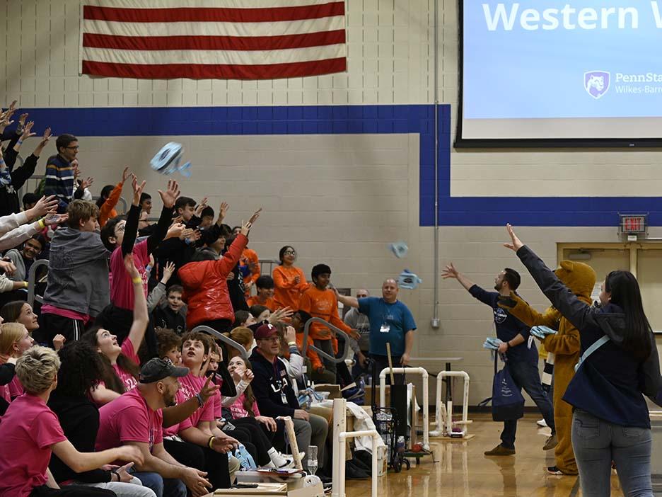 The Nittany Lion and several other people, at right, toss items to a crowd of people in the bleachers at left.