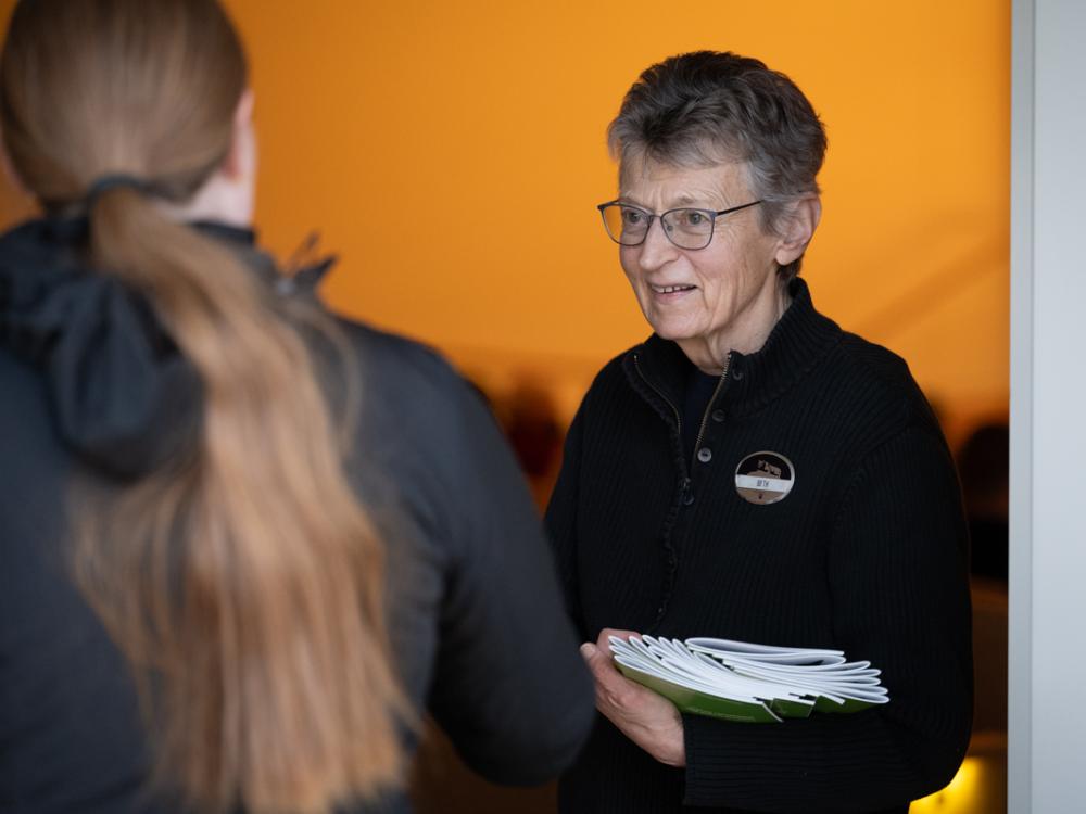 A woman wearing glasses holds a stack of programs that she hands out to patrons entering an auditorium.