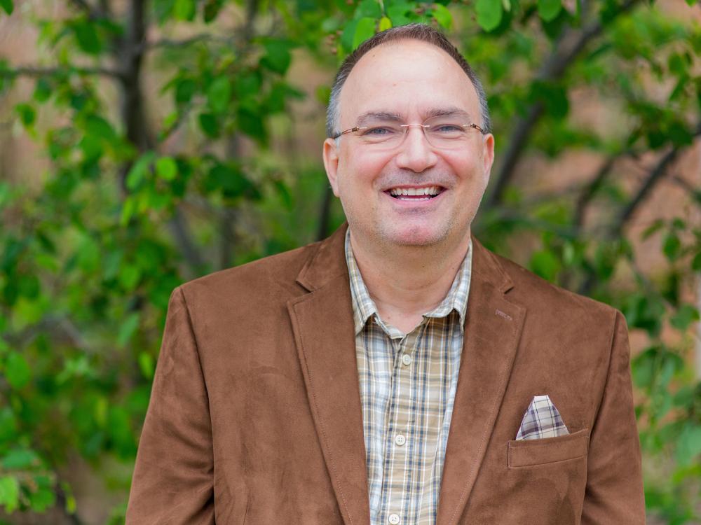 A photo of John Chapin wearing a brown jacket and smiling in a picture that was taken outdoors