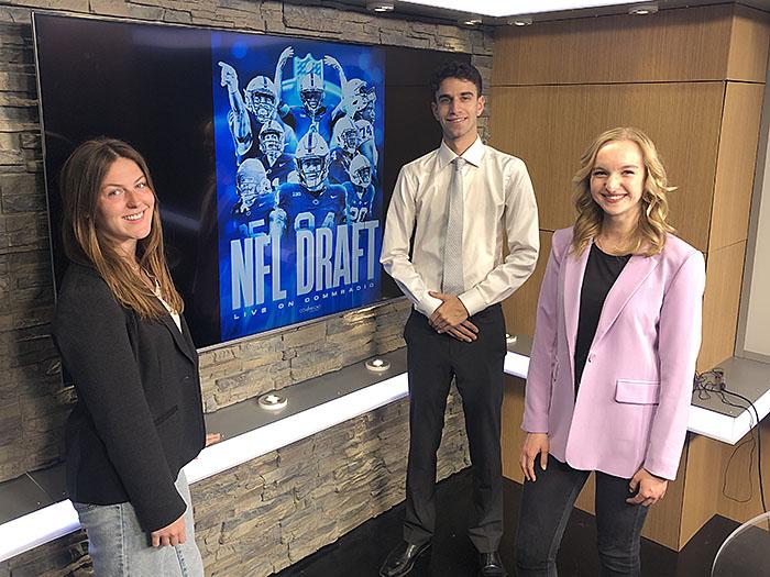 Three people stand in front of a television displaying "NFL DRAFT" graphic