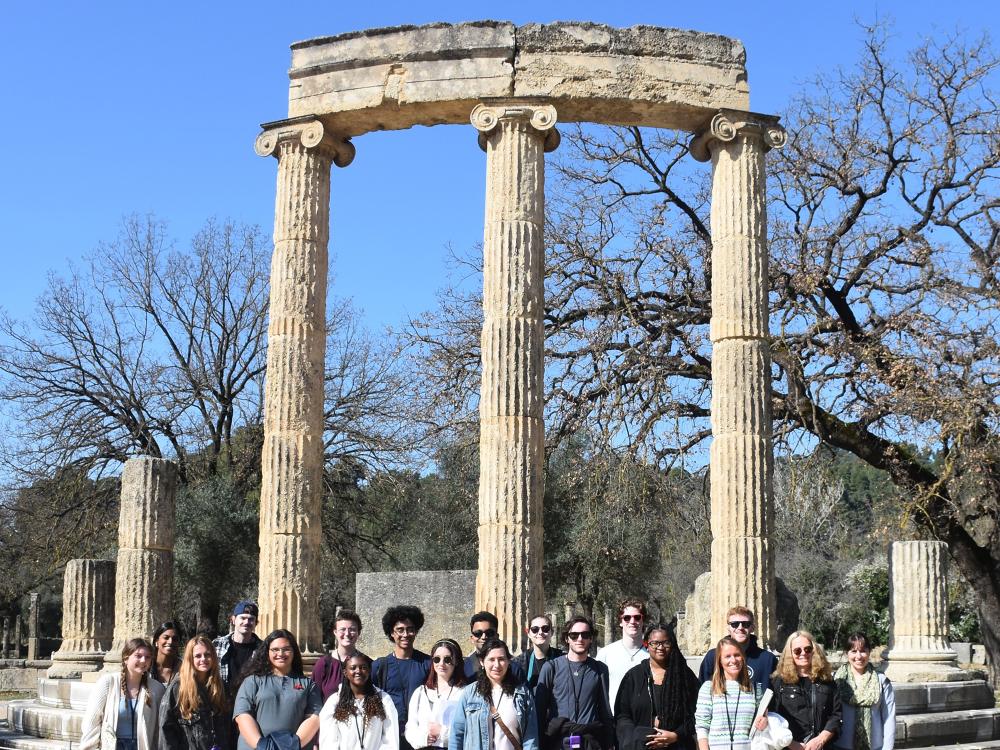 A group of students and faculty stands in front of one of the monuments at the site of Olympia in Greece.