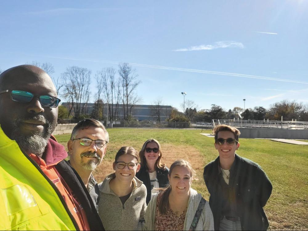 Six people take a selfie with a wastewater treatment plant in the background
