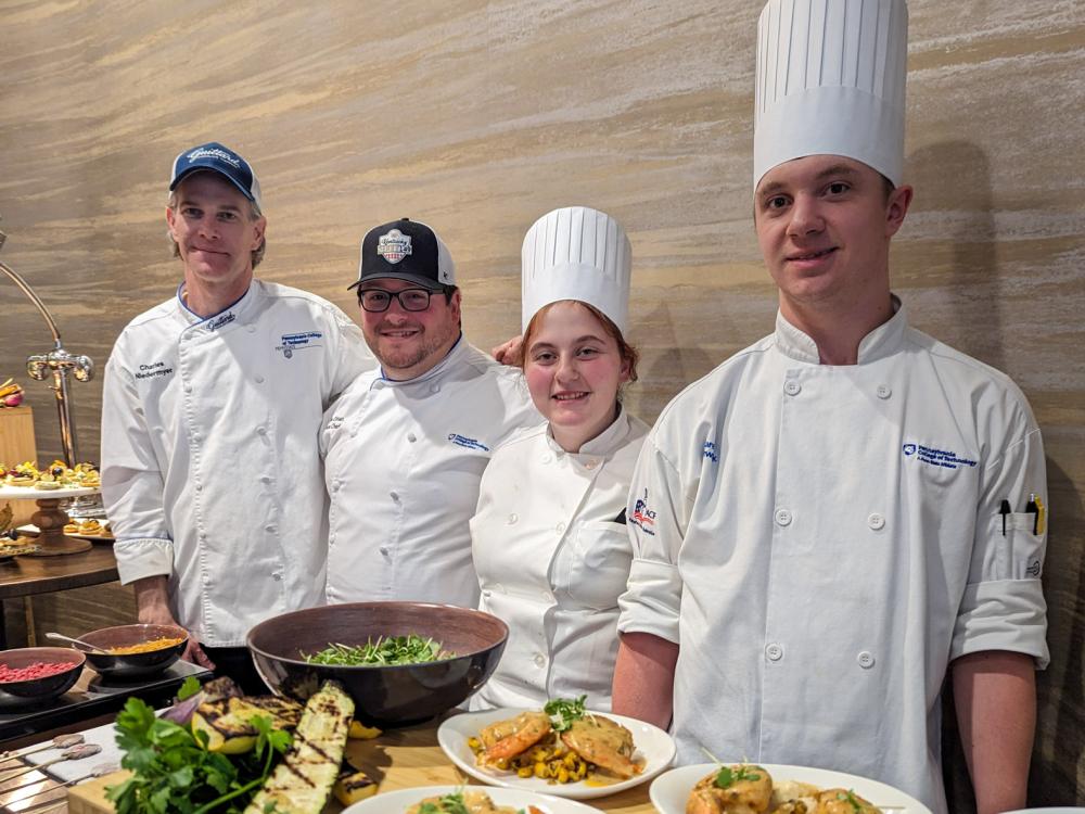 Four people wearing cooking attire stand with a table full of beautifully plated food