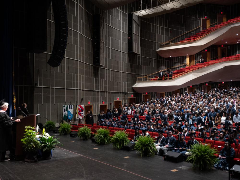 A master of ceremonies standing at a lecturn addresses a seated audience.