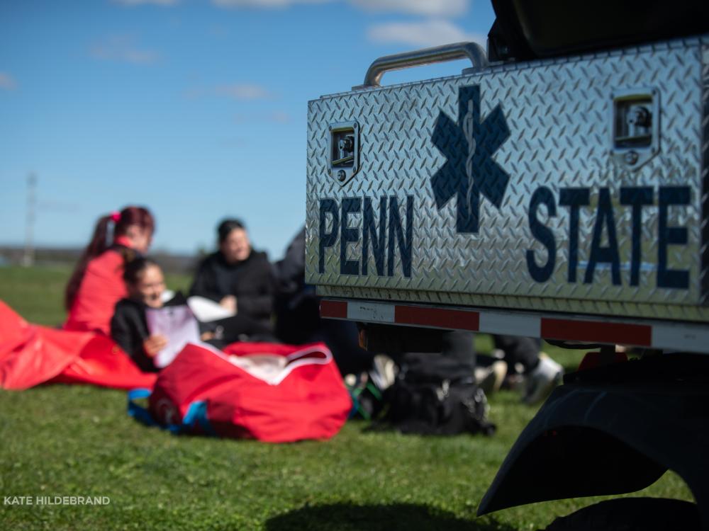 A Penn State emergency vehicle is parked on a grassy field with students sitting on the ground in the background.