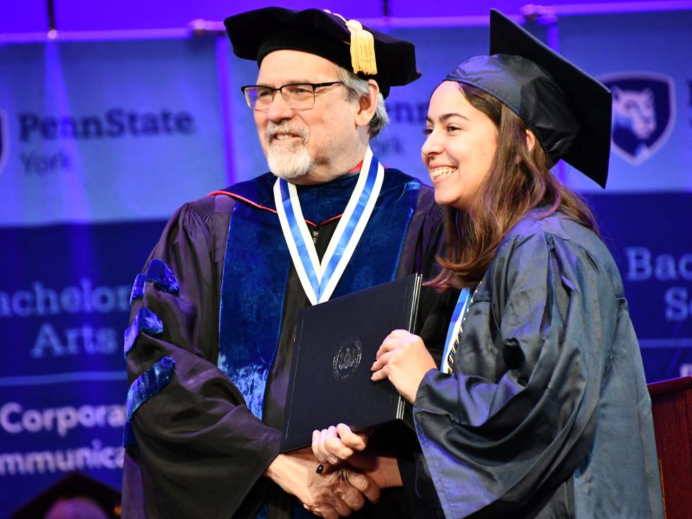 Man shakes hands with young lady accepting diploma, dressed in regalia. 