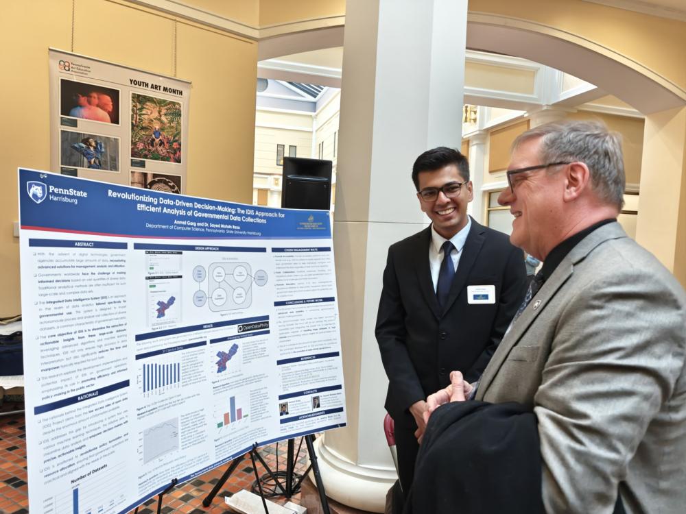 Anmol Garg stands next to his research poster in the state Capitol, while another person looks at the poster