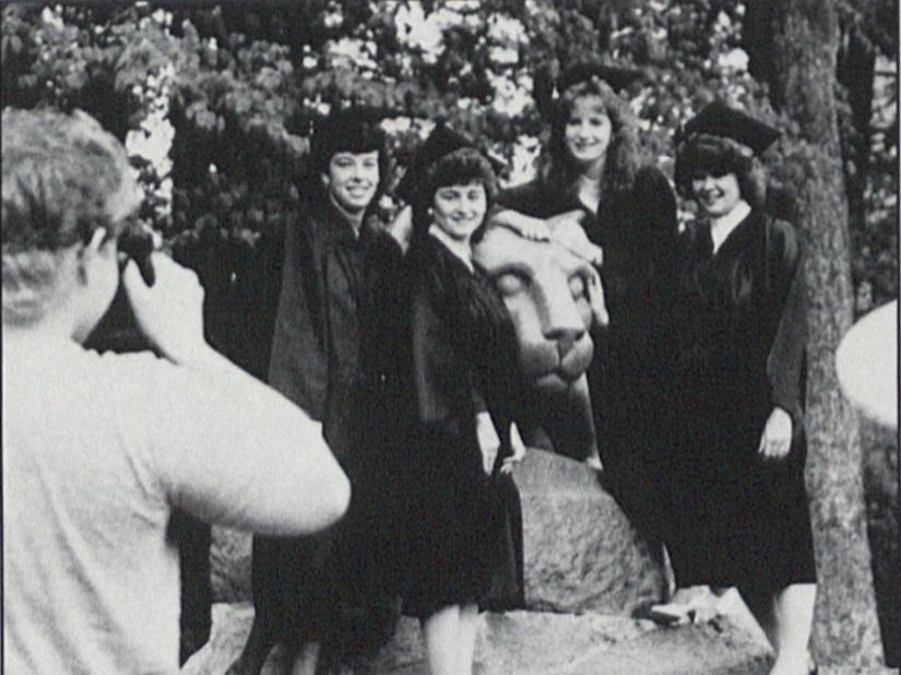 A group of four graduates pose for a camera in front of the Nittany Lion statue