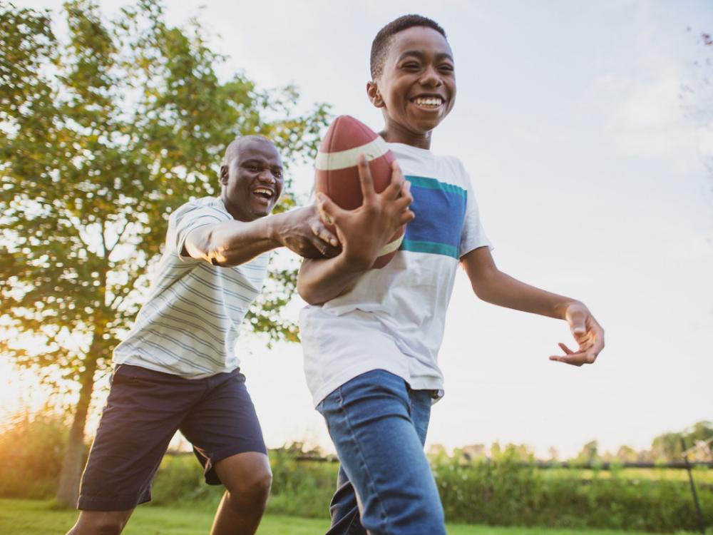 A boy and his dad laugh together while playing football on a lawn in the summertime.