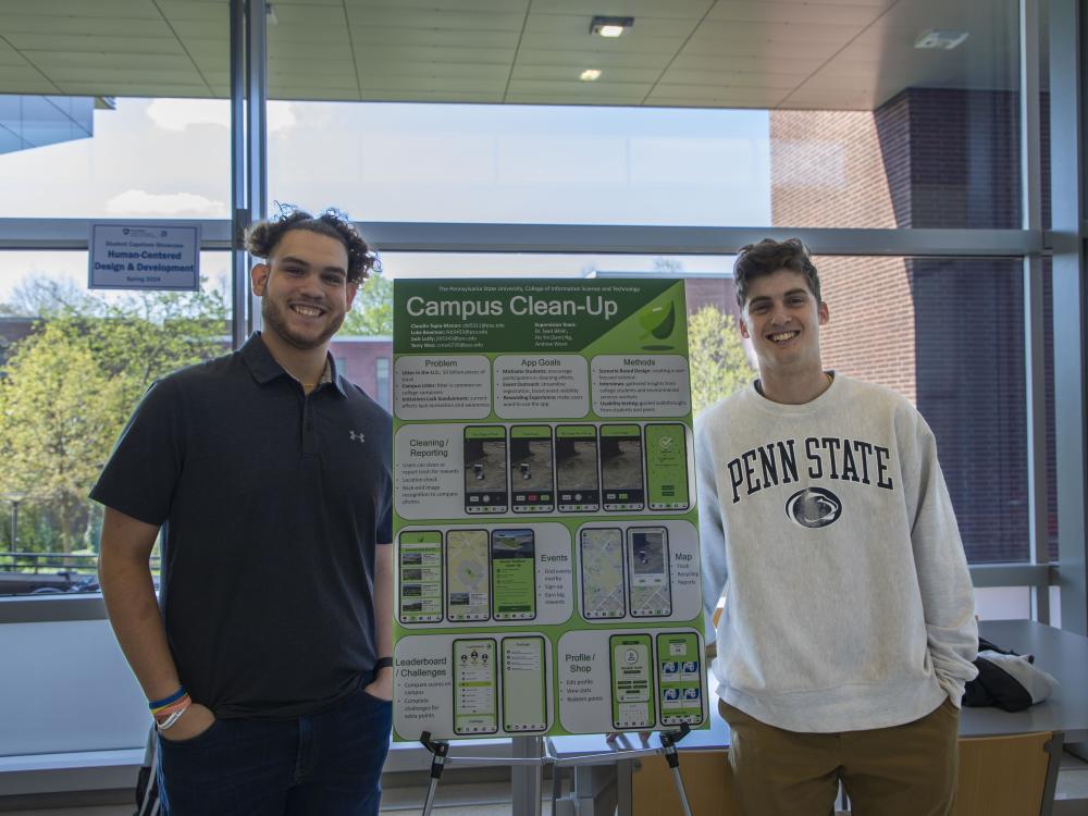 two smiling people stand next to academic poster in campus building