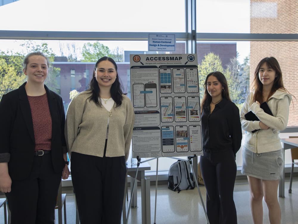 four college students stand next to poster in campus building