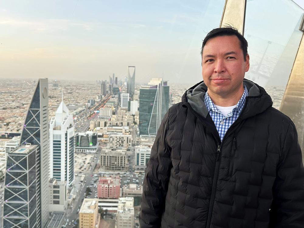 a man stands in front of a glass window showing a birds-eye view of Riyadh, Saudi ARabia, in the background