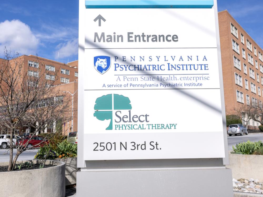 A brick, six-floor hospital is in the background. A sign in the foreground includes the logos for Pennsylvania Psychiatric Institute and Select Physical Therapy.