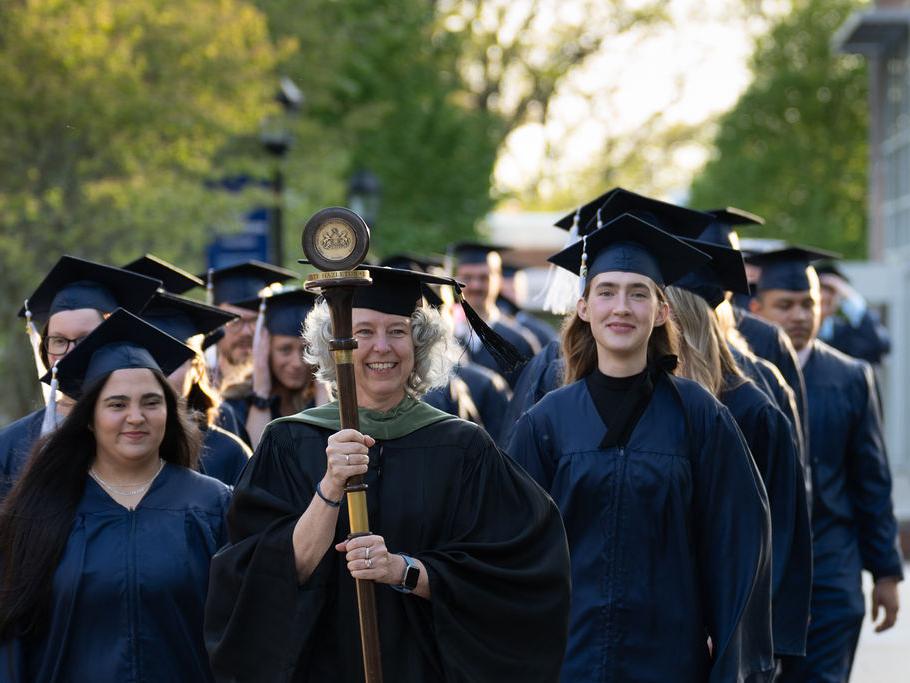 Graduates in caps and gowns in two rows walking down sidewalk led by faculty member in black robe carrying a staff and pendant.