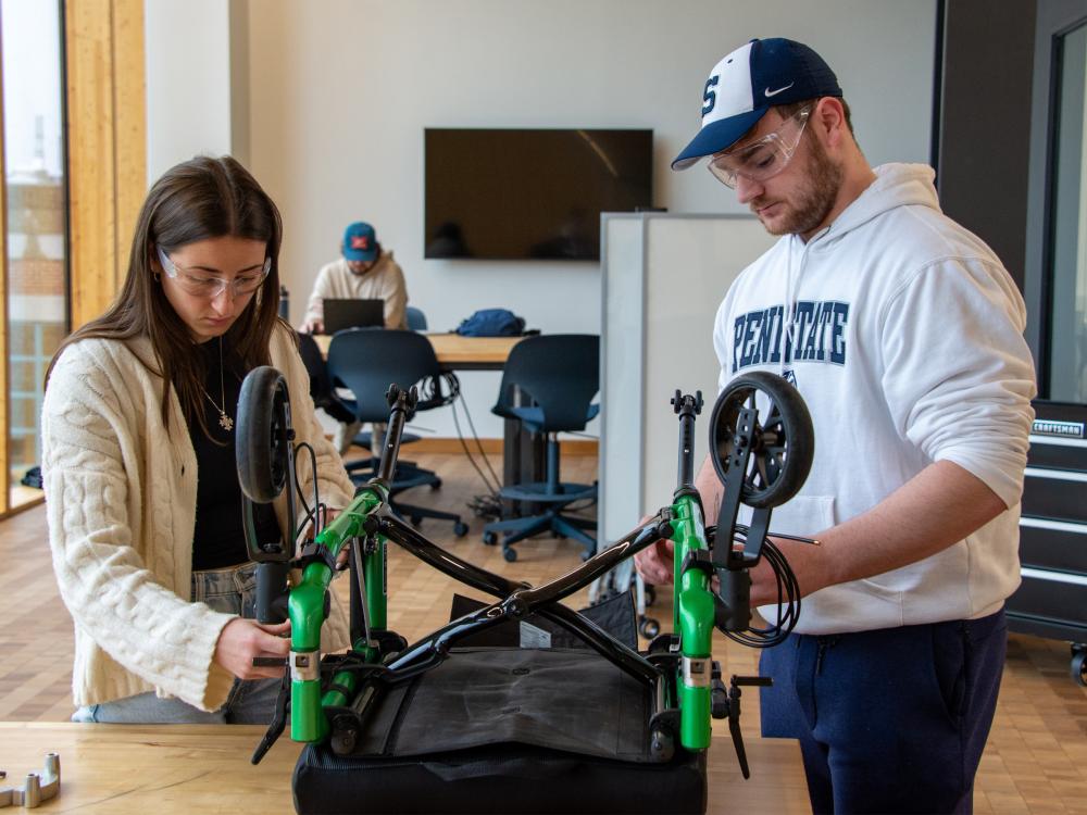 Two people wearing safety glasses adjust a wheelchair that is set upside-down on a table. 