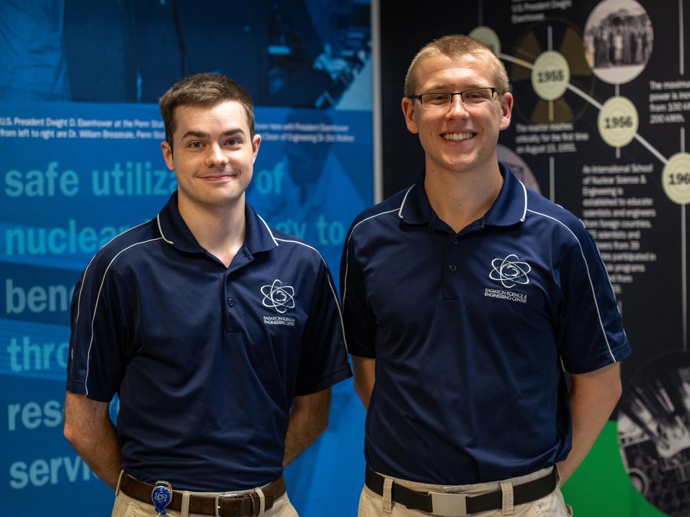 Two students wearing blue polos pose indoors behind a blue, black and green backdrop. 