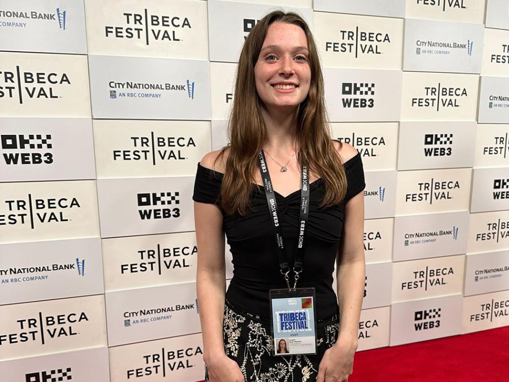 Long brown-haired girl in a black dress stands on a red carpet for a photo