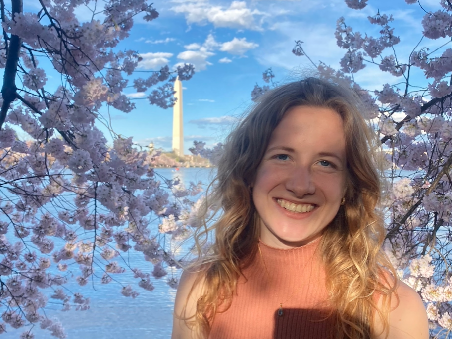 Chloe Connor stands among cherry blossoms with the Tidal Basin and the Washington Monument in the background.