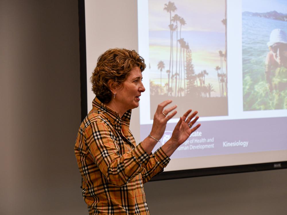 Woman giving a presentation at the front of a room, with large PowerPoint screen