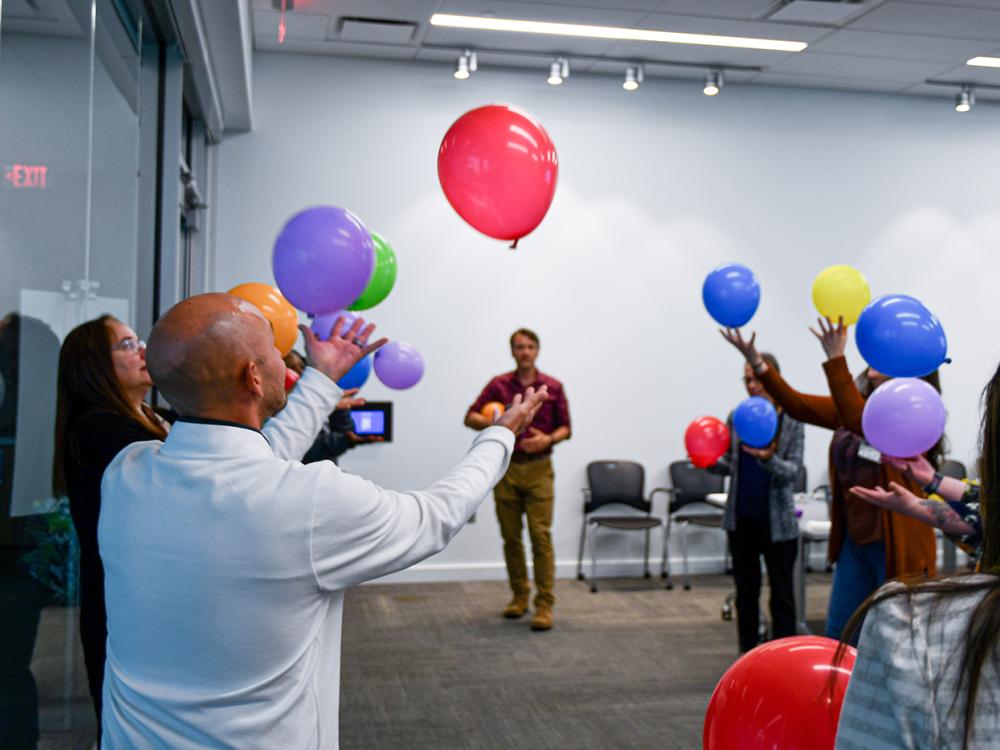 A group of people stand in a circle, tossing up colored balloons