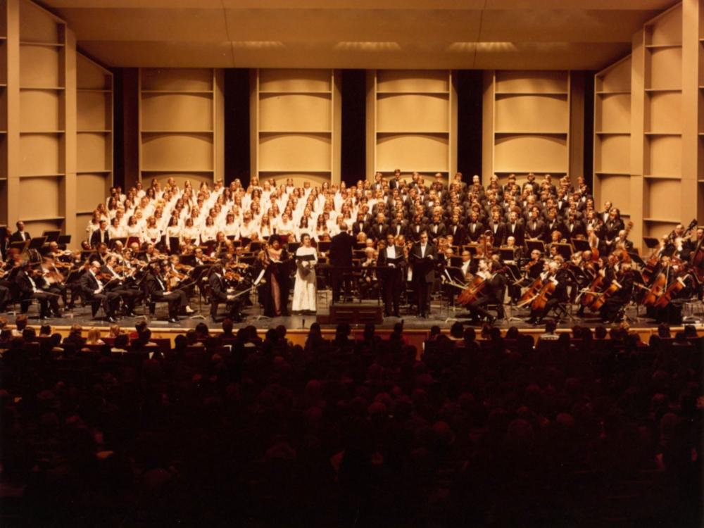 A number of choruses and classical musicians stand in formation on a stage.