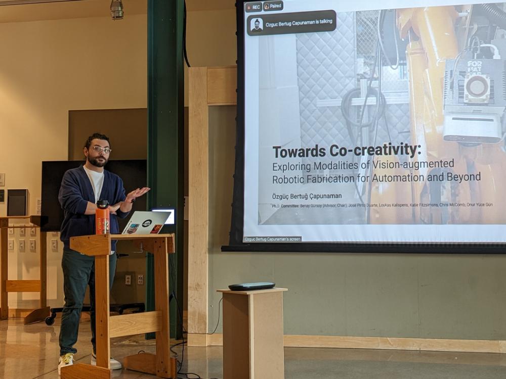 Özgüç Bertuğ Çapunaman stands at a podium delivering his thesis defense.