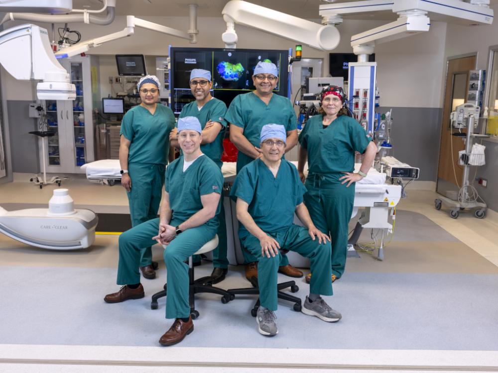 Six physicians in scrubs – four standing, two sitting – pose for a photo in a medical procedure room.