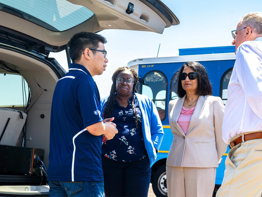 Four people talk in a group in front of a blue bus and next to a vehicle with an open truck. 