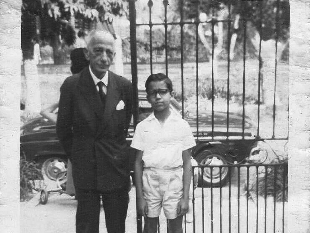 A black and white image of an adult and child posing for the camera in front of a gate and a car. 
