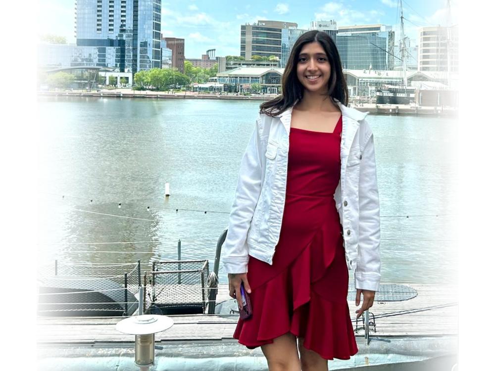 A person in a red dress and white jacket stands in front of a body of water with tall buildings in the background.
