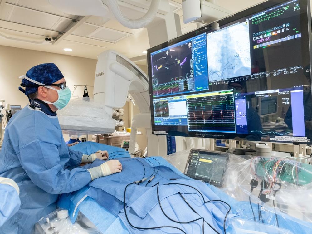 A physician in an operating room wearing scrubs and glasses stands over a patient and looks at a computer monitor.