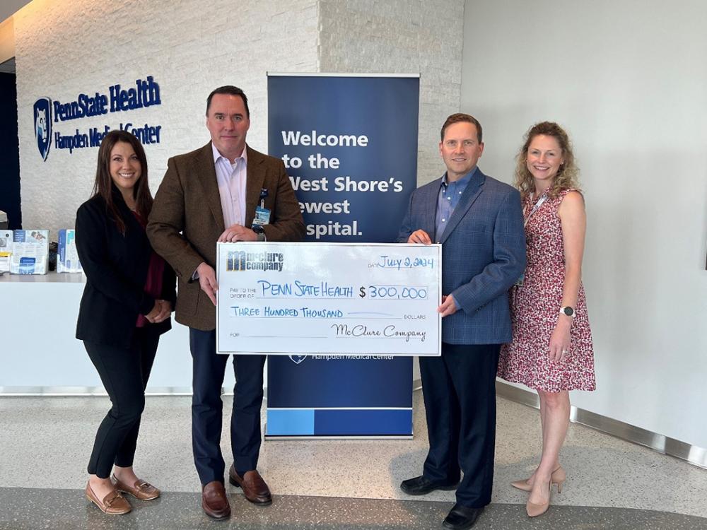 Two men and two women pose in front of a vertical banner that says, “Welcome to the West Shore’s newest hospital.” The gentlemen are in the middle holding a large presentation check for $300,000 written to Penn State Health from McClure Company.