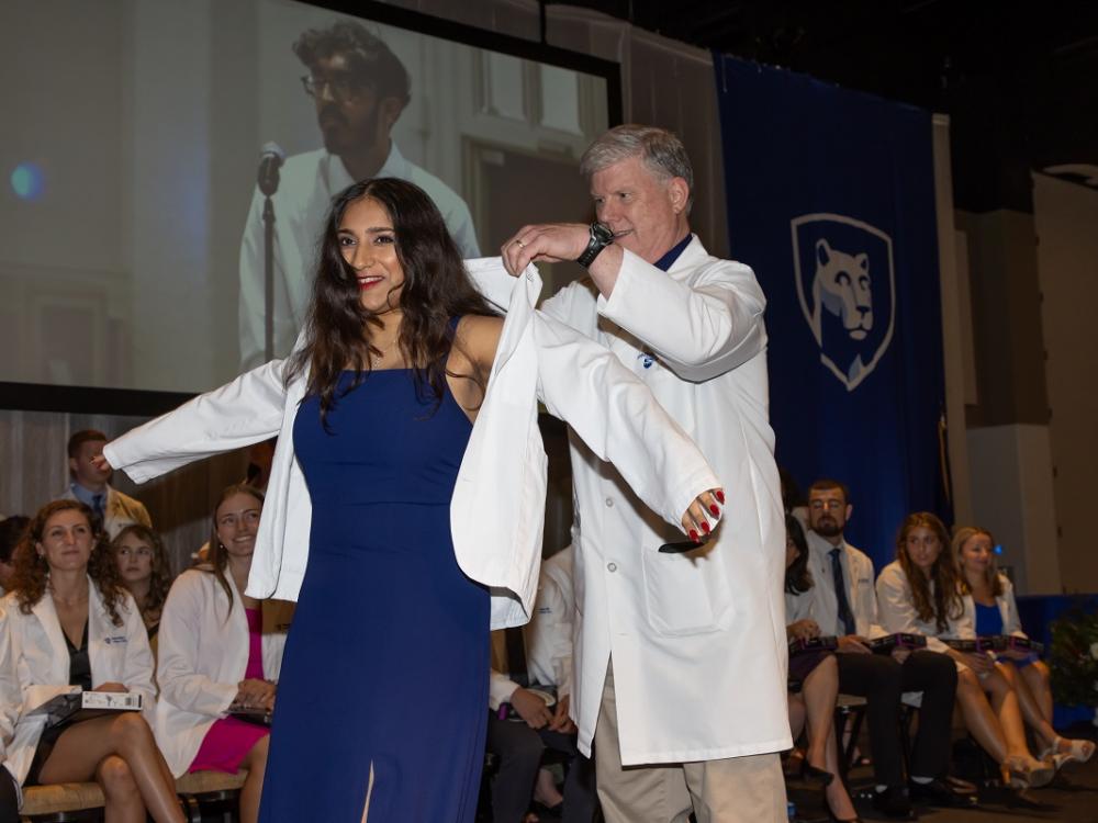 A man wearing a white doctor's coat presents a young woman with her own doctor's coat at the Penn State College of Medicine White Coat Ceremony