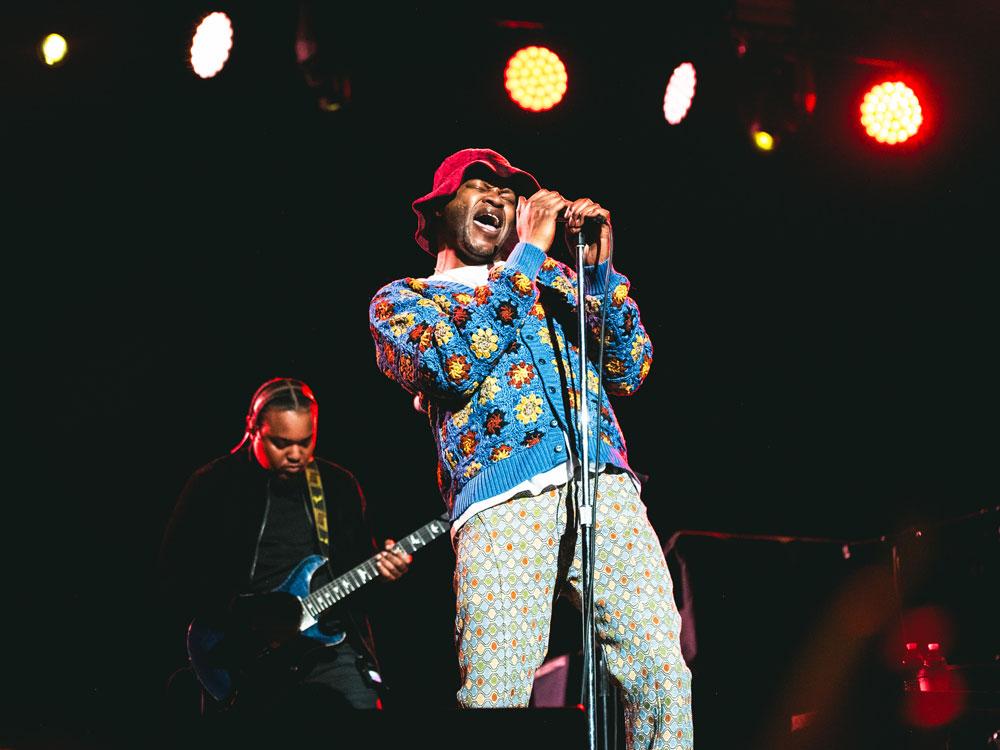 A man wearing a crocheted sweater and pants holds a microphone with both hands as he sings into it.