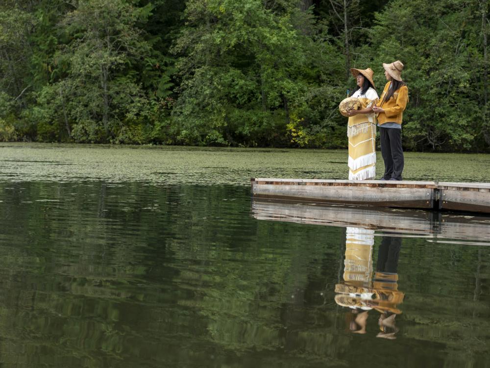 Two people stand on a dock near a lake