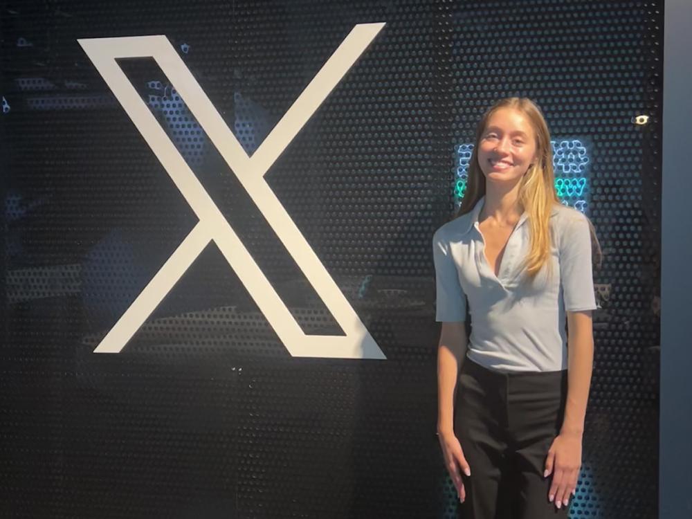 Girl with blond shoulder length hair stands in front of a logo for X